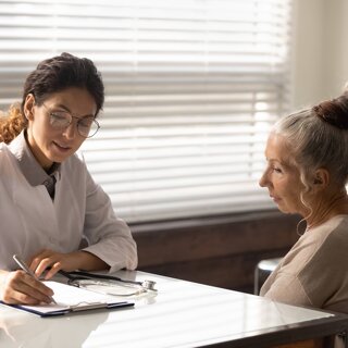 Médecin au travail avec des lunettes rédigeant un diagnostic des symptômes d'une patiente d'âge mûr dans un dossier médical.